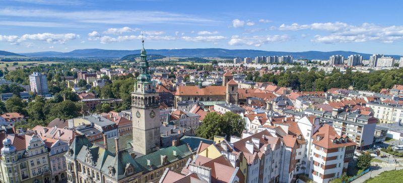 Widok ze szczytu góry na zabudowę Kłodzka z widocznymi szczytami gór w tle.