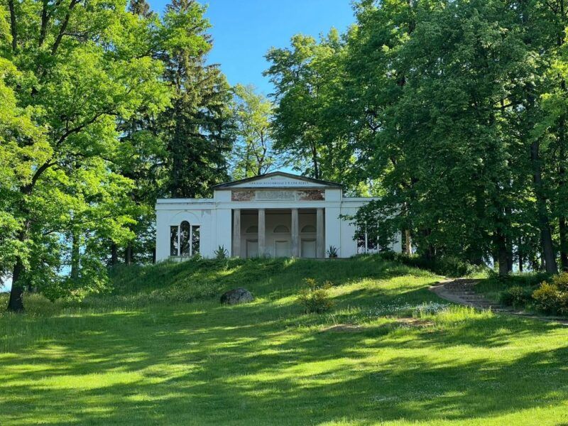 Biały budynek z portykiem znajdujący się na wzniesieniu wśród drzew.