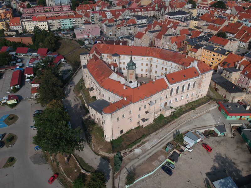 Zdjęcie bryły zamku, wykonane z drona. Widoczny jest niemalże trójkątny rzut obwarowań, łososiowy kolor fasady oraz czerwona dachówka.