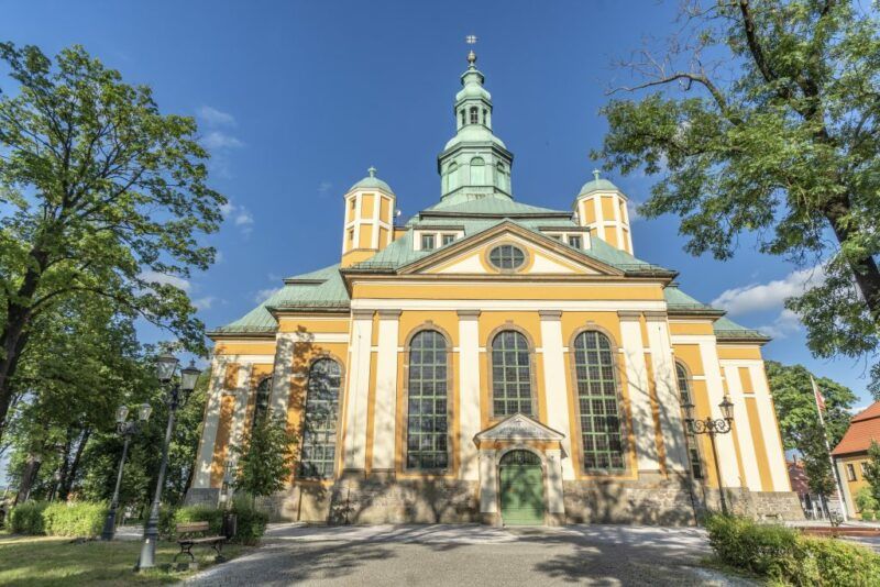 Widok na neogotycką budowlę z wysoką wieżą, ostrymi łukami i ozdobnymi detalami kamiennymi. Obok kilka drzew, na tle błękitnego nieba.