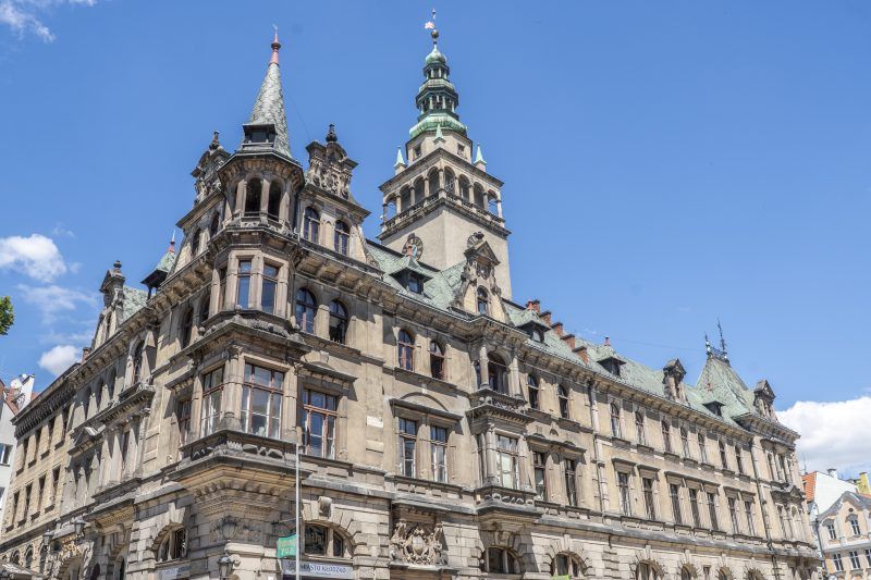 Widok na kłodzki ratusz, budowla w stylu neorenesansowym, wyróżniająca się smukłą wieżą zegarową oraz bogato zdobioną fasadą z detalami architektonicznymi.