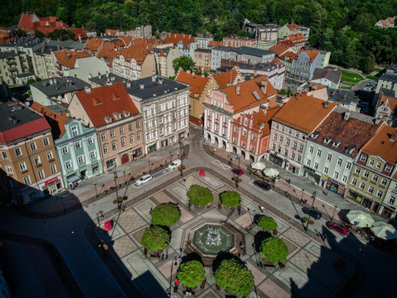 Zdjęcie z Drona przedstawiające stare zabytkowe kamienice, których pośrodku znajduje się zadbany plac z liczną zielenią, drzewami
