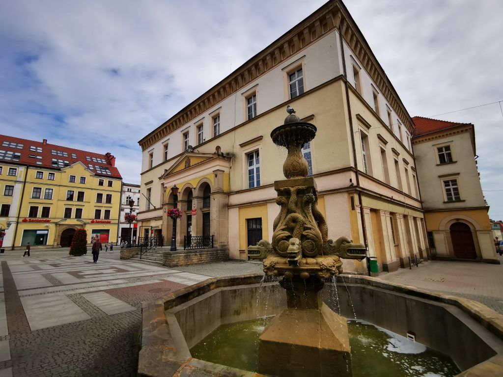 Widok na ratusz z biało-żółtą elewacją, w tle budynki, na pierwszym planie fontanna.