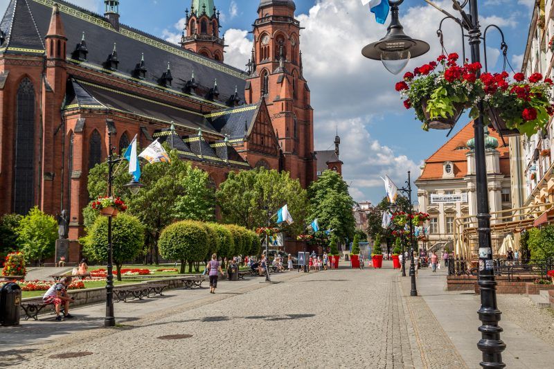 Zdjęcie przedstawia szeroki deptak w legnicy z ceglanym kościołem widocznym w tle