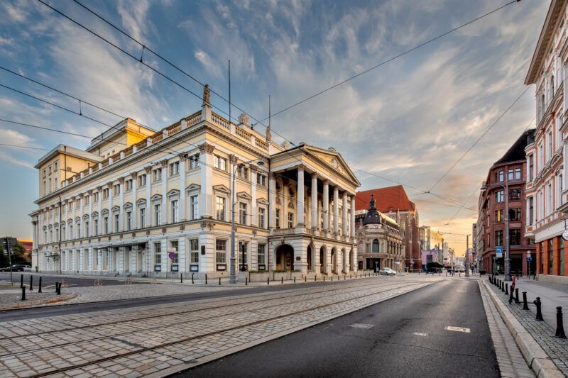 Widok z poziomu ulicy na duży klasycystyczny budynek opery z gotyckim kościołem i ulicą w tle
