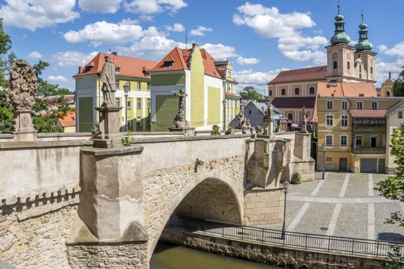 Słoneczny widok na zabytkowy most z figurami patronów, w tle dwuwierzowy kościół