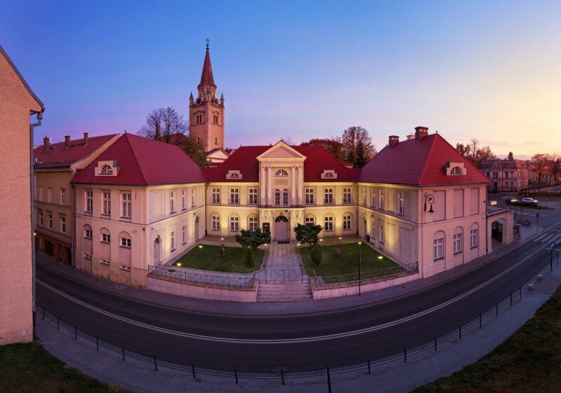 Zdjęcie z frontu na fasadę pałacu, w którym obecnie mieści się muzeum. W tle widoczna wieża. Zdjęcie robione na tle błękitnego, wieczornego nieba.