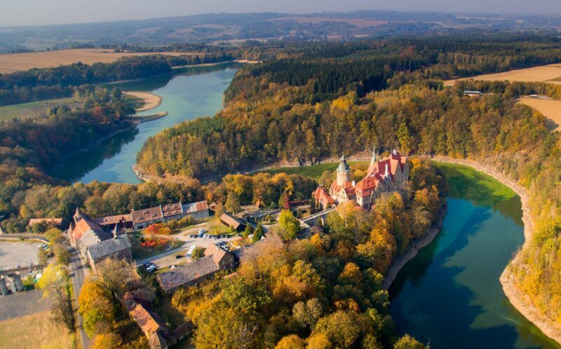 Widok z góry na zamek umiejscowiony nad brzegiem jeziora, które okala las w jesiennych barwach.