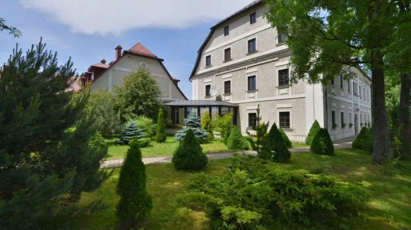 Obraz przedstawia trzypiętrowy, szaro biały budynek, będący tłem dla ogrodu pokrytego trawą, krzewami oraz tujami. Pomiędzy częścią roślin a domem znajduje się chodnik z kostki.