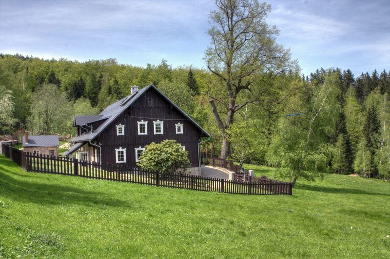 Zdjęcie przedstawia drewniany domek z białymi okiennicami, przysłonięty krzewem, otoczony drewnianym płotem na pokrytej trawą polanie. W tle widać las