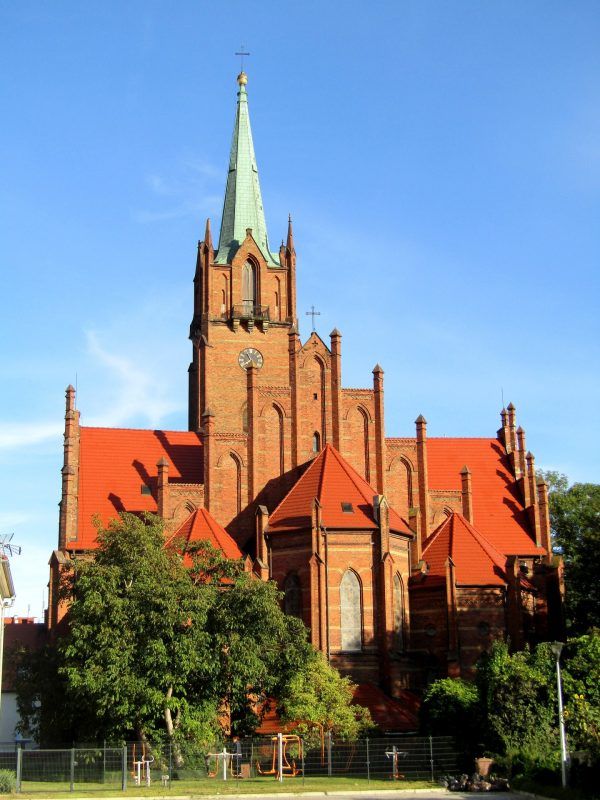 Zabytkowy kościół z czerwonej kostki i ze strzelistą wieżą otoczony drzewami