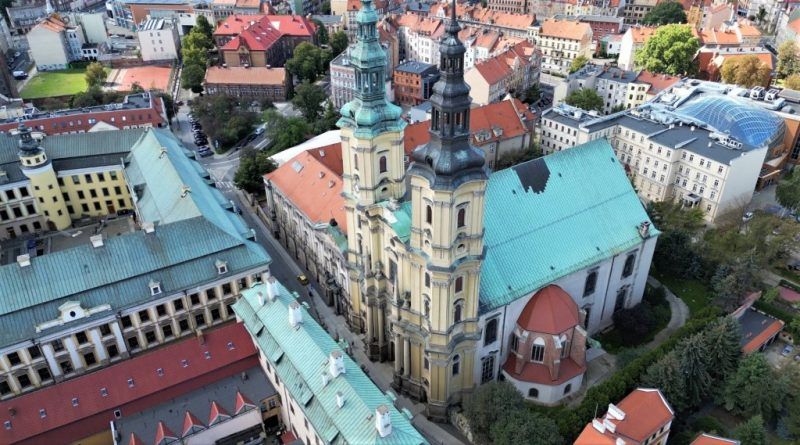 Barokowy kościół w zółtym kolorze z dwiema wieżami, obok znajdują się budynki klasztoru