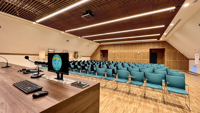 Nowoczesna sala konferencyjna z widokiem z perspektywy mównicy. Na pierwszym planie biurko z mikrofonami, klawiaturą i monitorem z logo Karkonoskiego Parku Narodowego. Przed biurkiem ustawione rzędy turkusowych krzeseł z składanymi blatami. Wnętrze wykończone zostało drewnem.