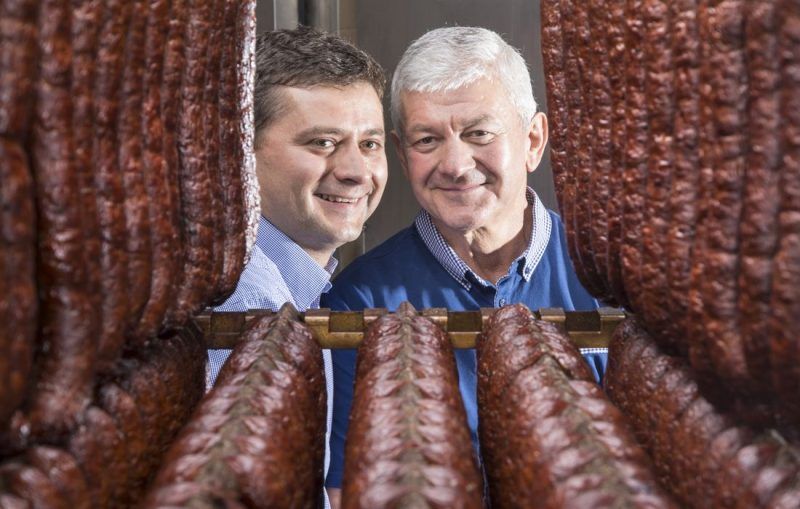 Dwóch uśmiechających się mężczyzn stojących pomiędzy zwisającymi pętami kiełbas.  
