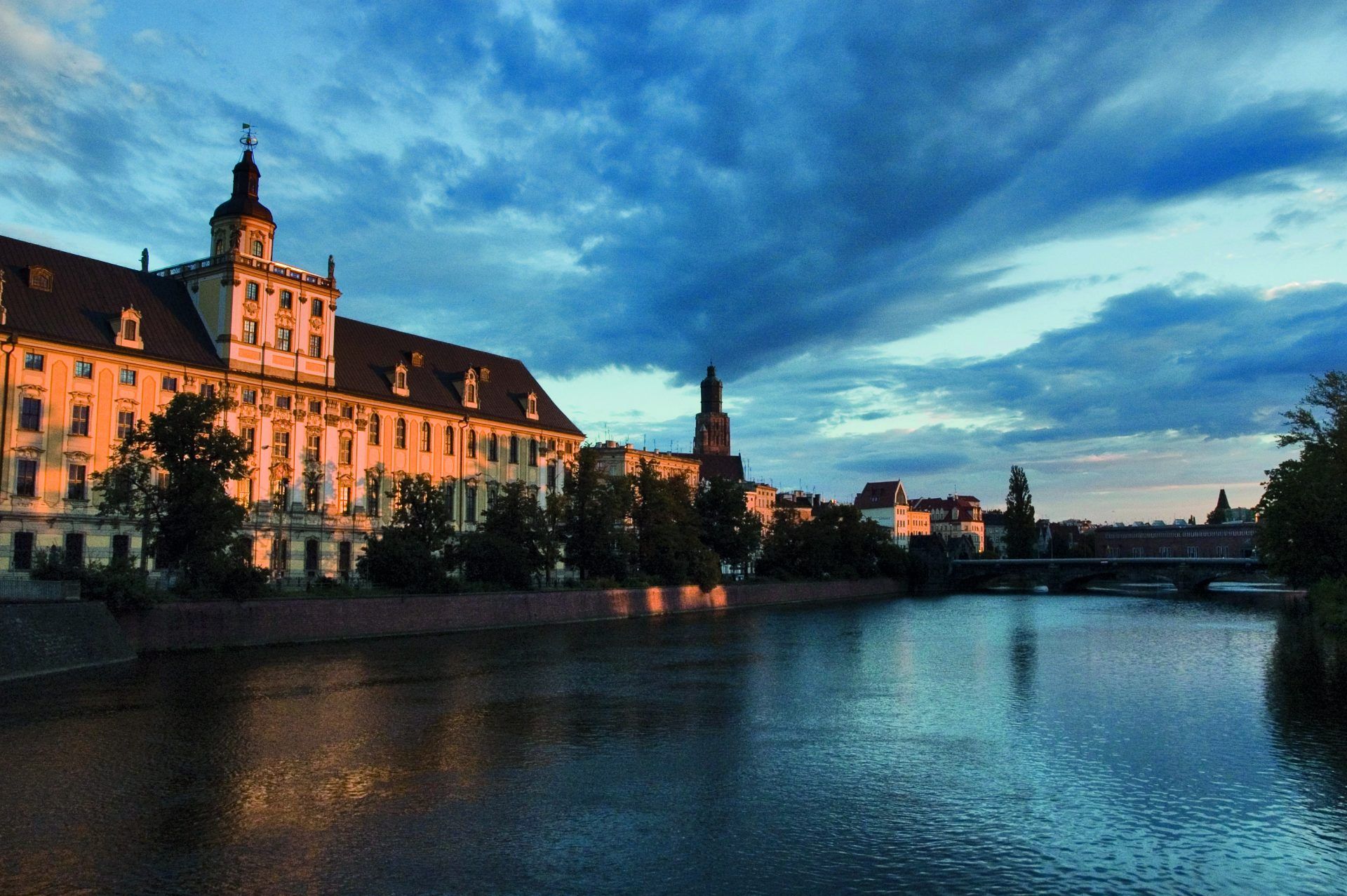 Zdjęcie przedstawia zabytkowy budynek uniwersytetu z wieżą oraz szeroką rzeką przepływającą obok budynku.