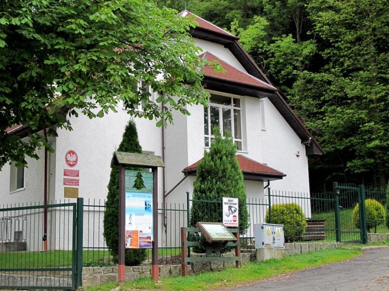 Na zdjęciu widać baił budynek zasłonięty z lewej strony koroną drzewa. Przed budynkiem stoi płot oraz dwie tablice informacyjne Karkonoskiego Parku Narodowego