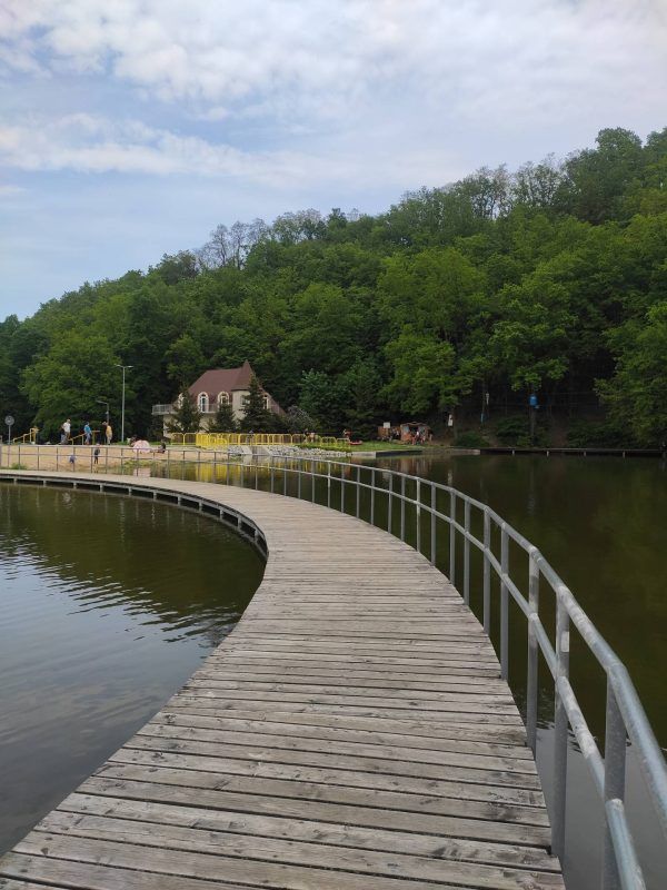 Zdjęcie przedstawia kładkę na zalewie w Złotoryi. Na drugim planie, na tle lasu, znajduje się mała plaża oraz budynek przysłonięty drzewami.