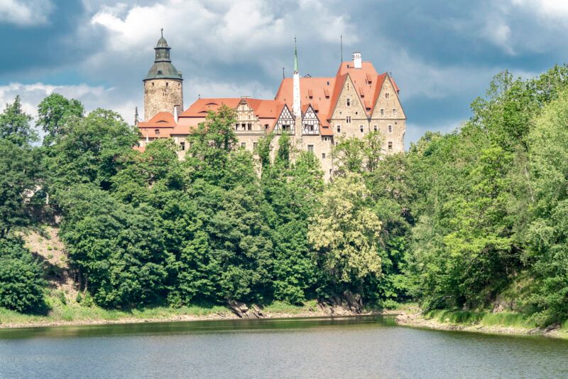Masywny zamek z okrągłą wieżą przysłonięty drzewami. Na pierwszym planie zbiornik wodny.
