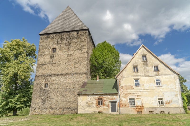Wieża zbudowana z jasnego kamienia, z czterospadowym dachem. Obok dwa mniejsze budynki, znacząco nowsze.