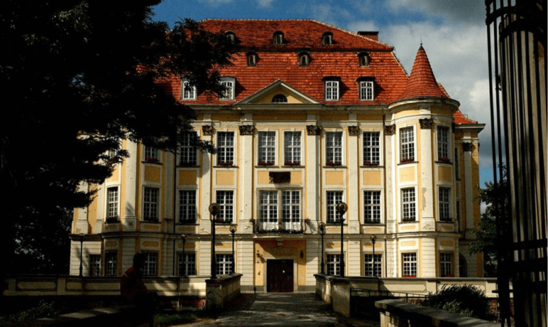 Zamek z czerwonym dachem, jasną elewacją oraz kwiatami na parapetach.