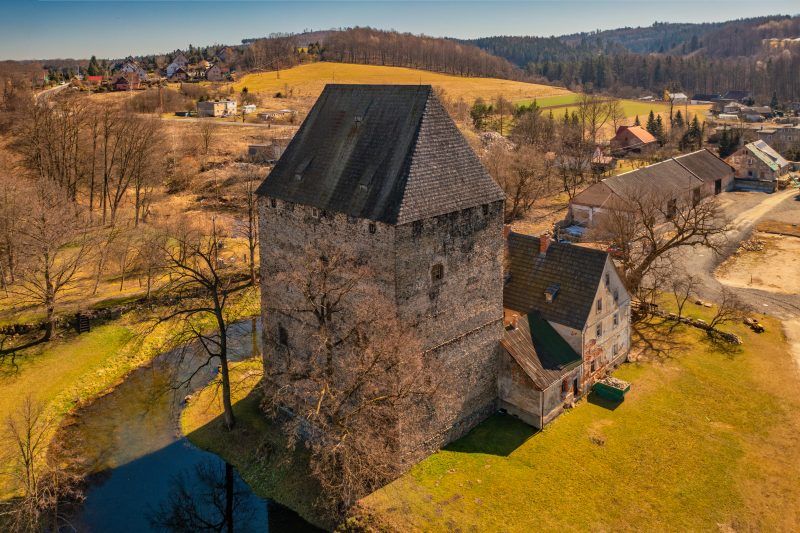Solidna, prostokątna wieża z kamienia w stylu gotyckim, z wysokim, spadzistym dachem, otoczona fosą i przylegającym budynkiem mieszkalnym.