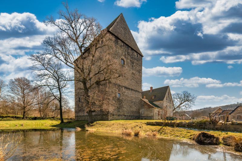Solidna, prostokątna wieża z kamienia w stylu gotyckim, z wysokim, spadzistym dachem, otoczona fosą i przylegającym budynkiem mieszkalnym.