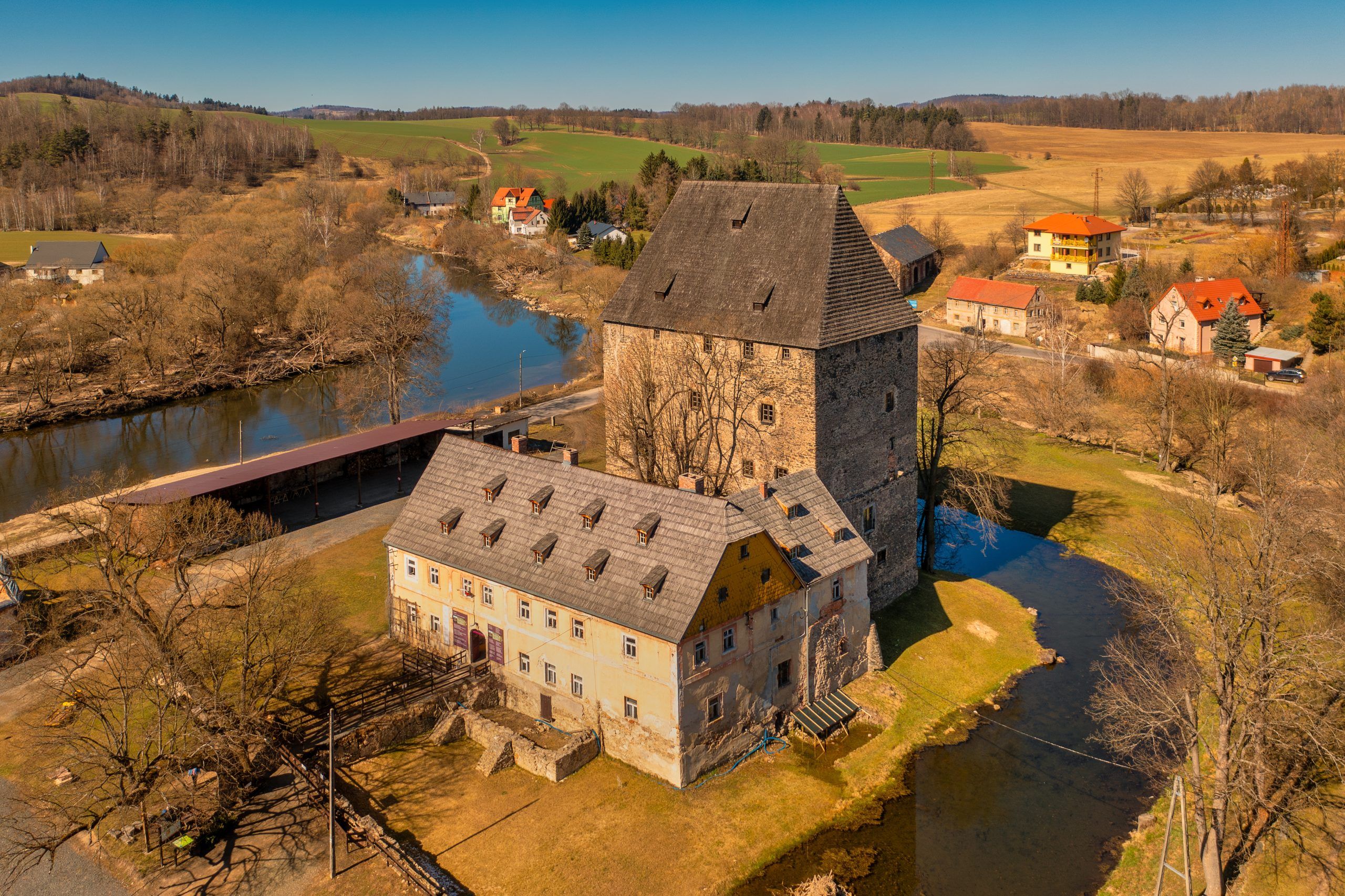 Solidna, prostokątna wieża z kamienia w stylu gotyckim, z wysokim, spadzistym dachem, otoczona fosą i przylegającym budynkiem mieszkalnym.