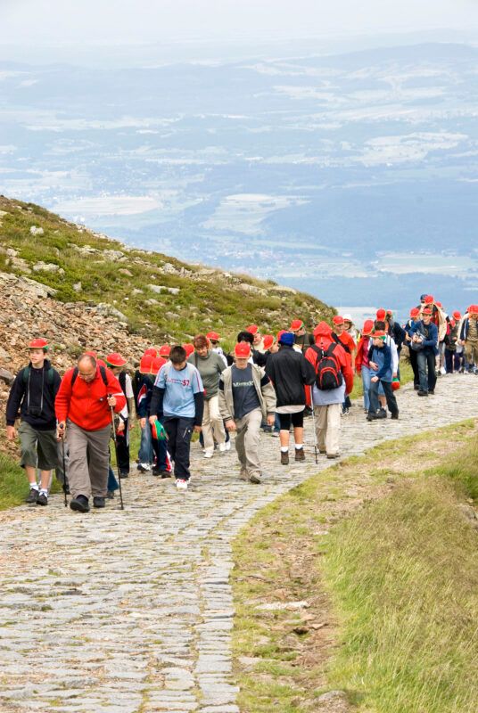 Duża grupa ludzi w czerwonych czapkach, podążająca za ubranym w czerwony polar przewodnikiem górskim, maszeruje po wybrukowanym szlaku. W tle widoczne zielone zbocze z kamieniami.