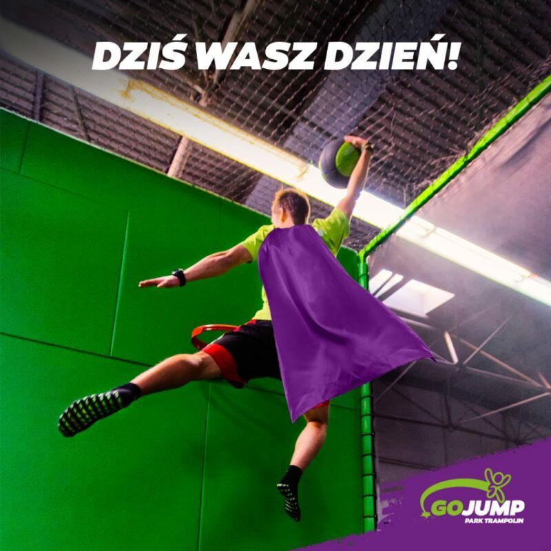 Mężczyzna z fioletową peleryną podskakuje z trampoliny do zawieszonego kosza by wrzucić piłkę