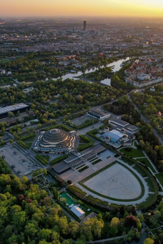 Widok z lotu ptaka na budynek Hali Stulecia oraz miasto Wrocław. W tle zachód słońca.