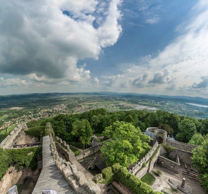 Widok znad murów kamiennego zamku otoczonego gęstym lasem. W tle ciekawa panorama miasta oraz najbliższej okolicy.