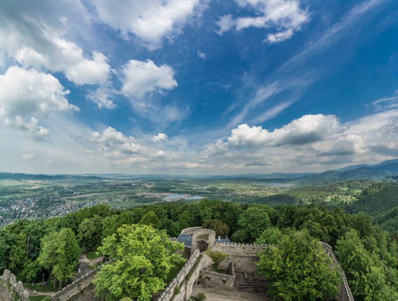 Widok znad murów kamiennego zamku otoczonego gęstym lasem. W tle ciekawa panorama najbliższej okolicy ze wzgórzami łąkami, stawem.