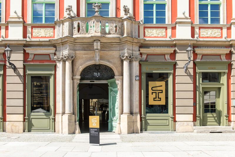 Na zdjęciu widnieje wrocławska kamienica z dużymi oknami, zdobieniami i pomarańczowymi akcentami. Widać również logo Muzeum Pana Tadeusza. 