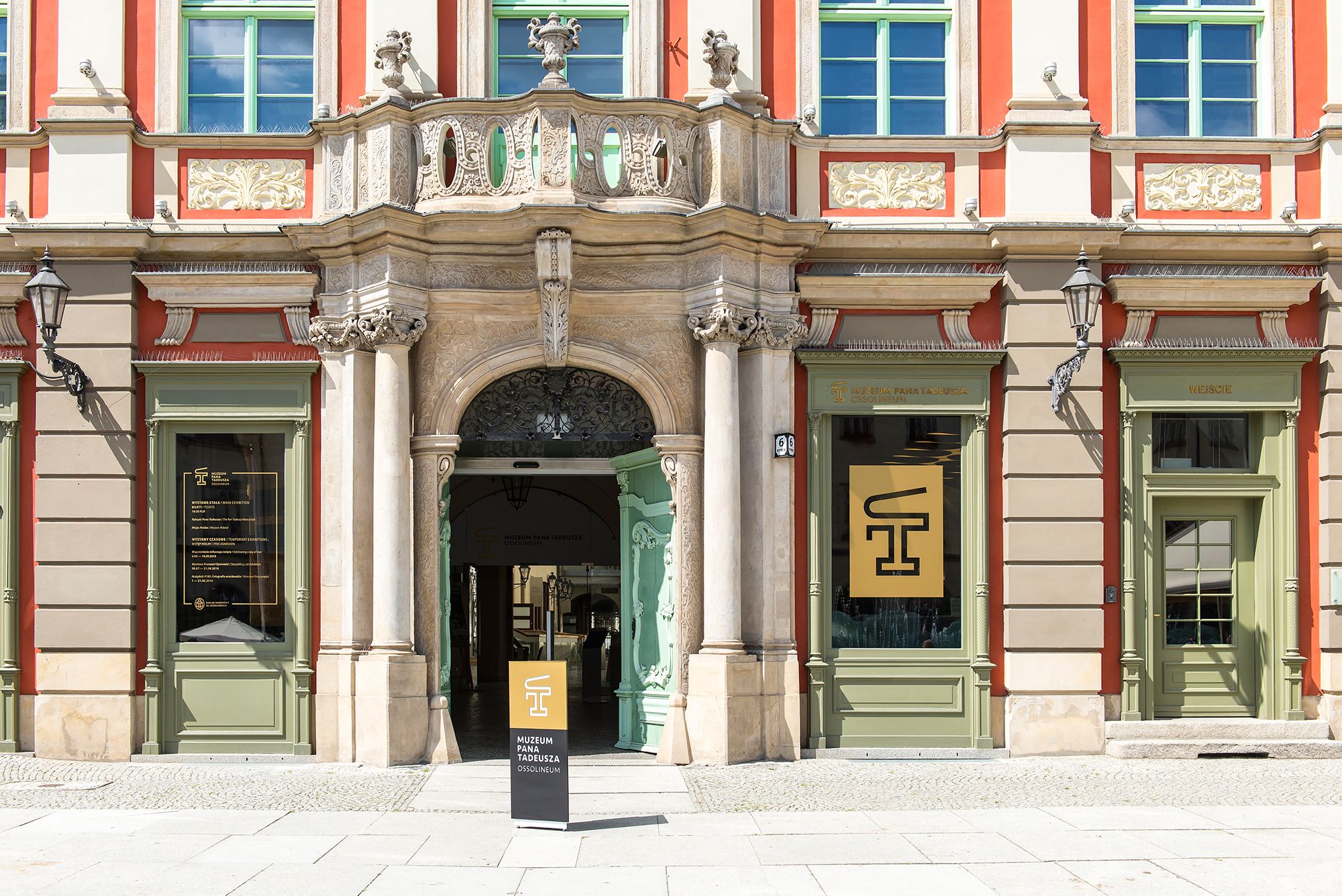 Na zdjęciu widnieje wrocławska kamienica z dużymi oknami, zdobieniami i pomarańczowymi akcentami. Widać również logo Muzeum Pana Tadeusza. 
