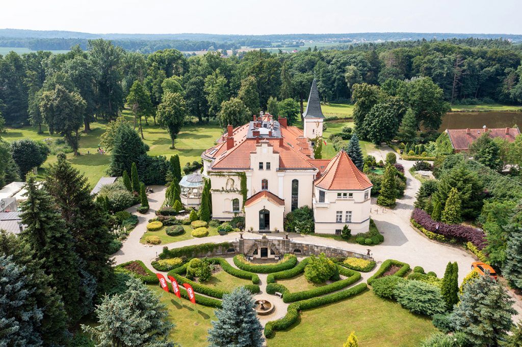 Widok na pałac z lotu ptaka, jasna elewacja, czerwony dach, przed pałacem piękny zadbany ogród.