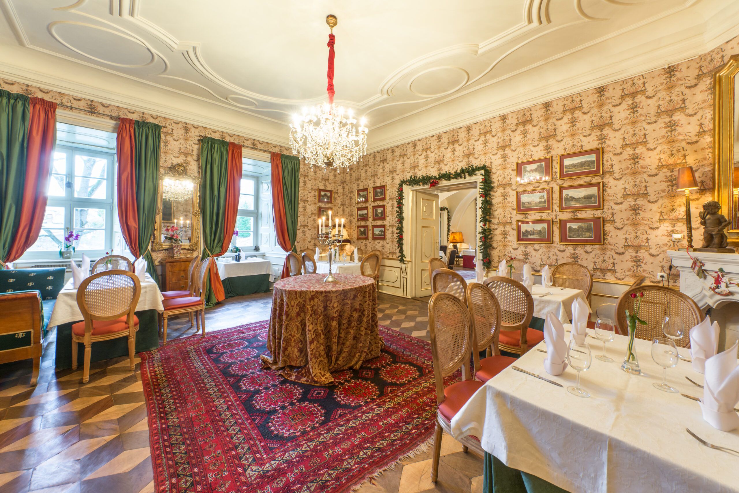 Wnętrze restauracji w stylu historyzującym, Na oknach duże, kotarowe zasłony. Stoły zastawione, przyszykowane na uroczystość. Na ścianach wzorzyste tapety oraz dużo zdjęć w ramach. Drewniany parkiet, przykryty prostokątnym dywanem we wzór geometryczny.