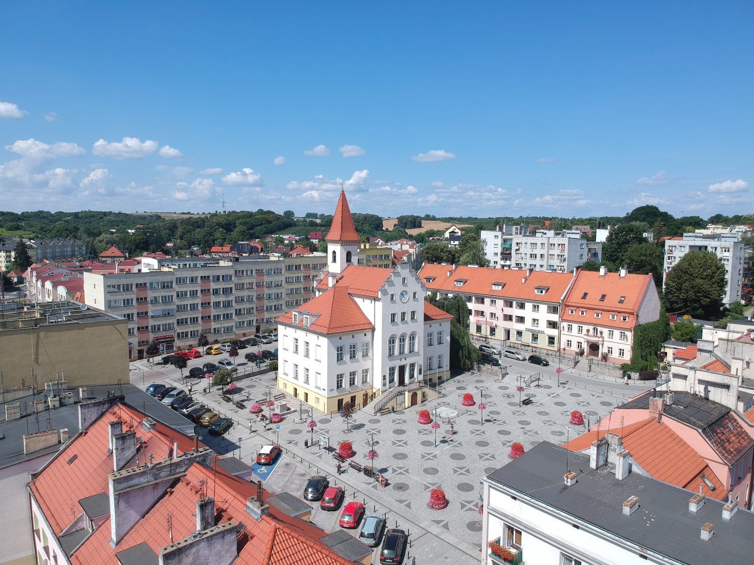 Zdjęcie przedstawia rynek Trzebnicy z centralnie ustawionym ratuszem z wysoką wieżą i czerwonym dachem. Przy głównym placu widać ustawione samochody.