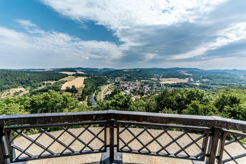 Panorama okolicy zamku widoczna z jego wieży. Wokół sporo zieleni, pola, rzeka, wzgórza. Na pierwszym planie widnieje barierka.