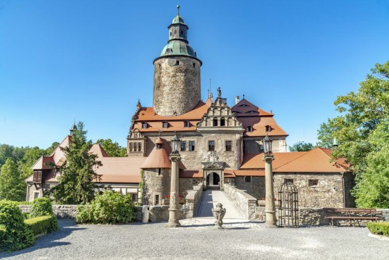 Kamienny zamek i prowadzący do niego most z wysoką okrągłą wieżą na tle błękitnego nieba.