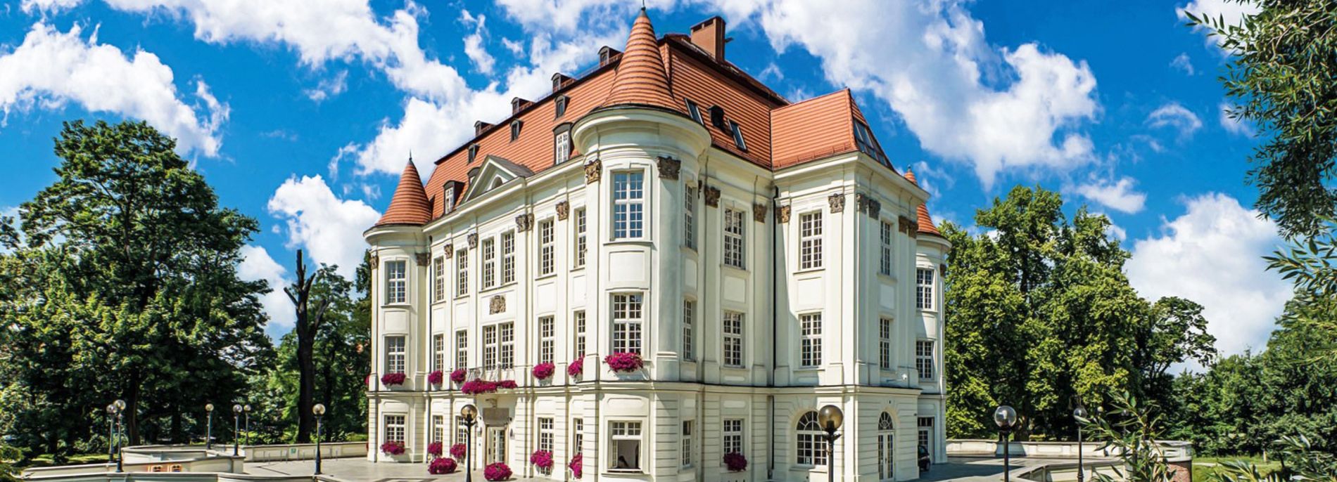 Zamek z czerwonym dachem, jasną elewacją oraz kwiatami na parapetach.