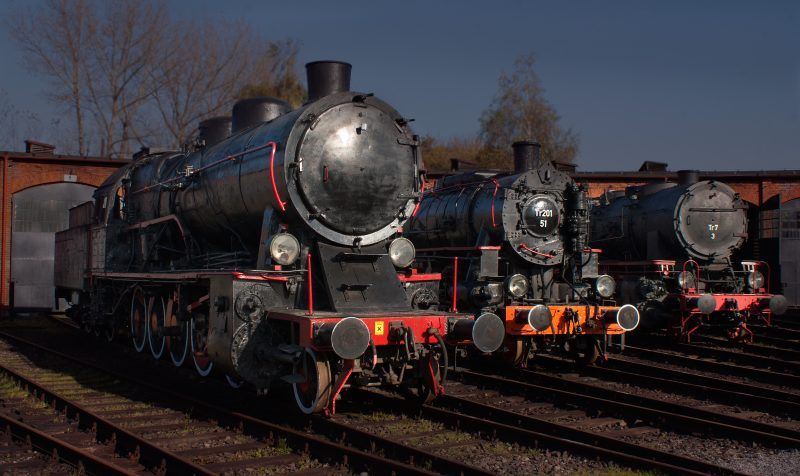 Trzy czarne lokomotywy  widoczne od frontu oświetlone promieniami słonecznymi