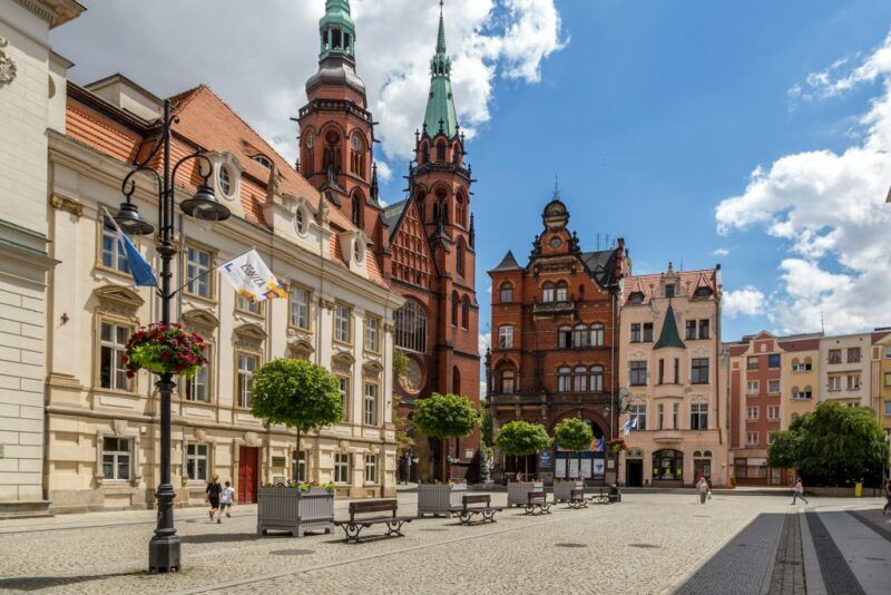 Zdjęcie przedstawia rynek i widoczne na nim kamienice oraz małe drzewka. Na drugim planie znajduje się ceglany kościół z dwiema wieżami.