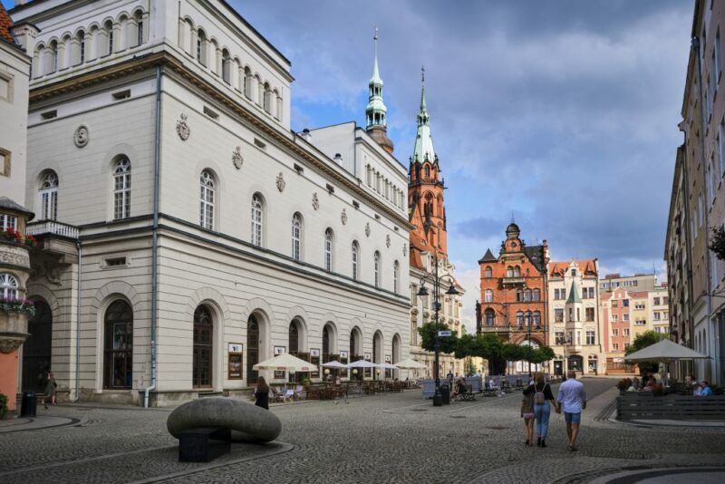 Wysoki budynek teatru w Legnicy z  kostkową płytą Rynku, w tle  ceglane wieże gotyckie katedry 