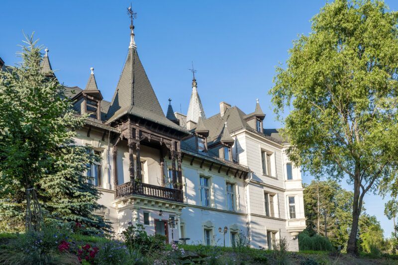 Oddalony widok na balkon na piętrze pałacu, wśród drzew.
