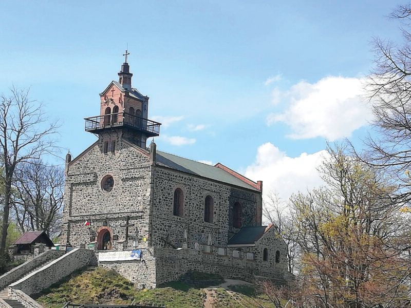 Widok na kamienny, nieotynkowany kościół z dwuspadowym dachem i niewielką wieżą w centralnej części fasady na tle nieba.