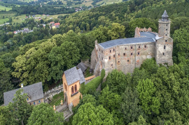 Majestatyczne ruiny gotyckiego zamku, położone na wzniesieniu, w górzystym krajobrazie, wybijające się pośród zieleni drzew. 