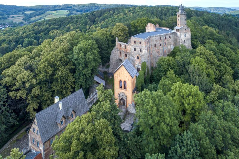 Majestatyczne ruiny gotyckiego zamku, położone na wzniesieniu, w górzystym krajobrazie, wybijające się pośród zieleni drzew.