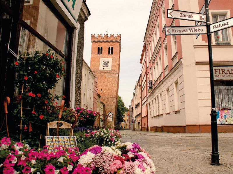 Widok z poziomu ulicy na krzywą, ceglaną nieotynkowaną wieżę. 