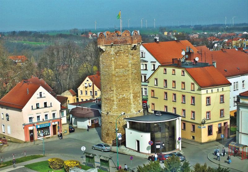 Widok z lotu ptaka na kamienną, średniowieczną basztę wybudowaną na planie koła z widocznymi wiatrakami w oddali.