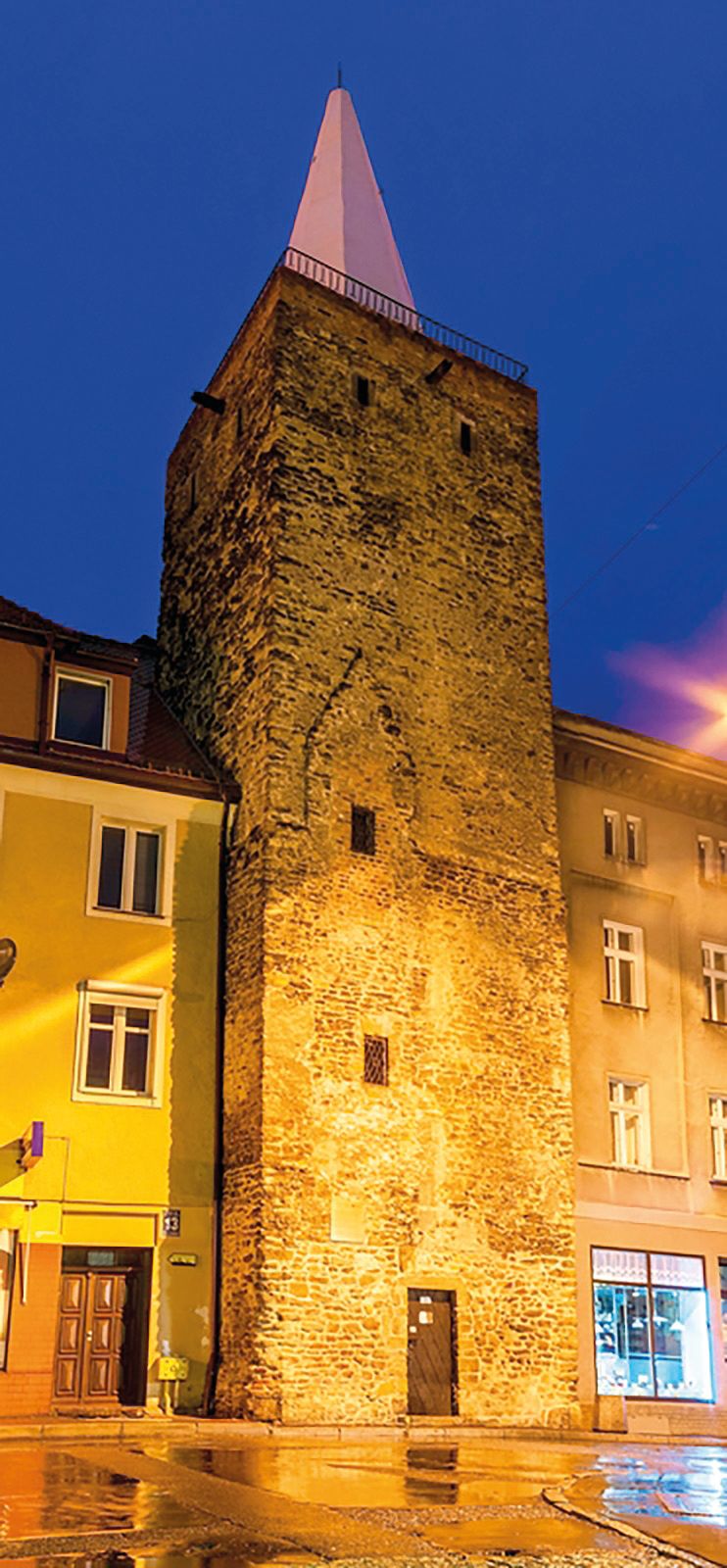 Nocny widok na średniowieczną, kamienną wieżę, kwadratową w swym planie, z przylegającymi do niej budynkami mieszkalnymi.
